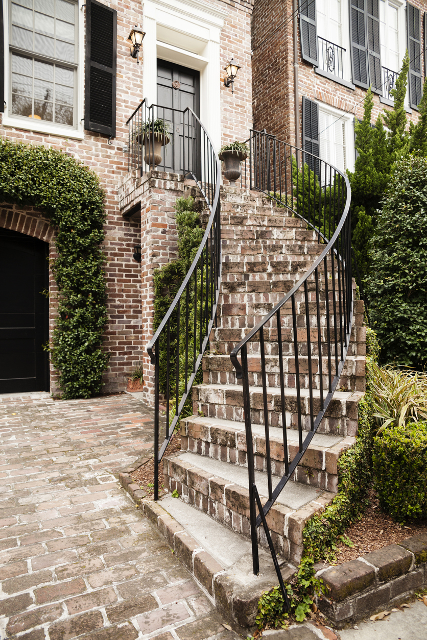 A beautiful, historic building in Charleston, South Carolina, USA. It has a curved, brick staircase with wrought iron railings leading from the brick driveway up to the front door. Ivy lines the garage, and lush foliage is all along the staircase.